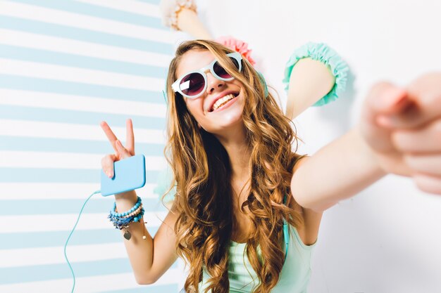 Ritratto del primo piano della ragazza beata in occhiali da sole e braccialetti alla moda in posa con il segno di pace. Affascinante giovane donna con i capelli lunghi che fa selfie tenendo il telefono e ascoltando la canzone preferita.