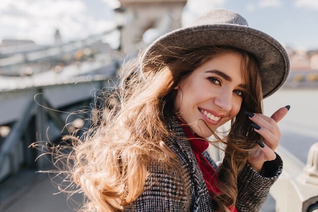 Ritratto del primo piano della ragazza bianca estatica indossa orecchini carini e cappello grigio alla moda