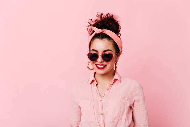 Ritratto del primo piano della ragazza con le labbra rosse e il panino in occhiali da sole. La donna in fascia rosa e camicia di cotone sorride su uno spazio isolato.