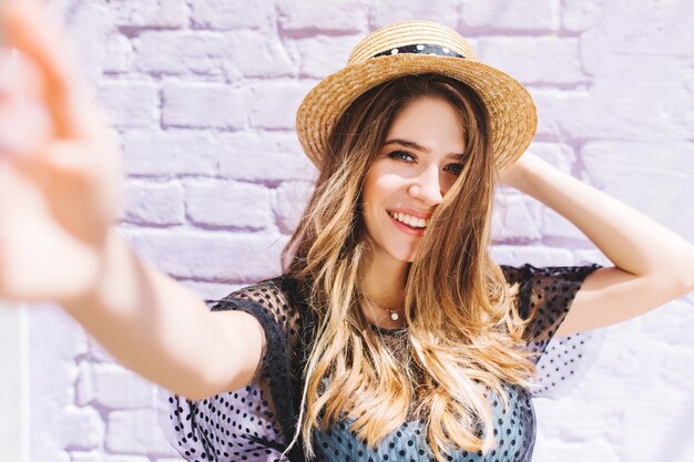 Ritratto del primo piano della ragazza felice con capelli di lunghezza medio-posteriore che fa selfie e che tiene il cappello di paglia