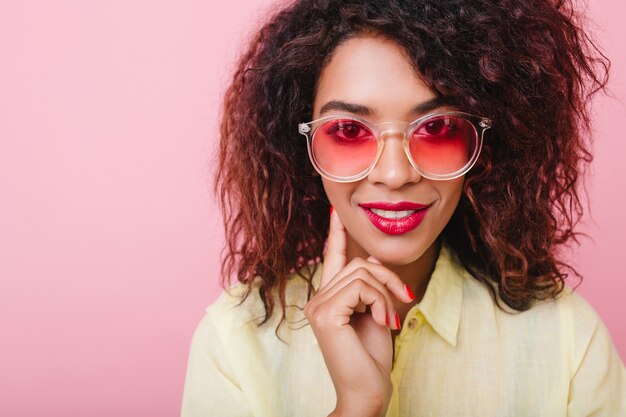 Ritratto del primo piano della ragazza interessata sensuale in occhiali rosa che tocca la guancia con la mano. Adorabile signora con pelle marrone e trucco luminoso sorridente.