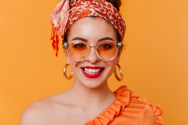 Ritratto del primo piano della signora alla moda con rossetto rosso, sorriso bianco come la neve sullo spazio arancione. Donna in velo e orecchini massicci che guarda l'obbiettivo.