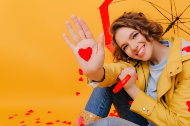 Ritratto del primo piano della signora di risata che tiene ombrello e cuore rosso di carta. Studio shot di bruna ragazza pallida sorridente durante il servizio fotografico nel giorno di San Valentino.