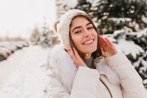 Ritratto del primo piano della splendida donna con gli occhi azzurri in posa sulla strada in un giorno di inverno nevoso. Foto all'aperto dell'affascinante modello femminile nella risata del cappello lavorato a maglia