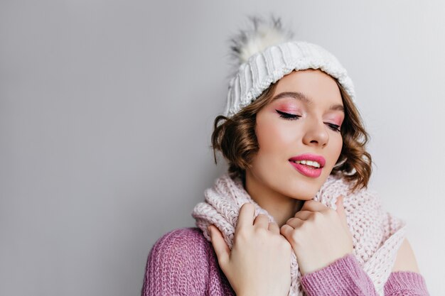 Ritratto del primo piano della splendida ragazza con trucco rosa brillante. Foto di interni di raffinata giovane donna con un grazioso cappello bianco e una sciarpa lavorata a maglia.