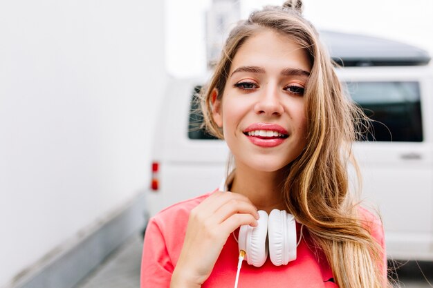 Ritratto del primo piano di adorabile ragazza bionda che cammina per strada in grandi auricolari bianchi