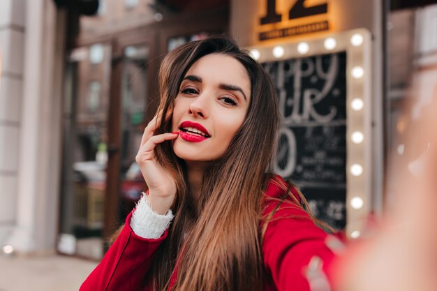 Ritratto del primo piano di bella donna con i capelli scuri che fa selfie sullo spazio della città della sfuocatura