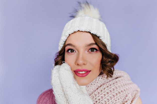 Ritratto del primo piano di bella donna europea in guanti di lana bianchi in posa sulla parete viola. Foto di interni di bella signora con il trucco rosa indossa un grazioso cappello lavorato a maglia.