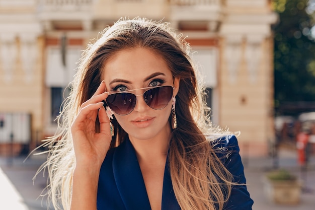 Ritratto del primo piano di bella donna vestita in giacca blu alla moda che cammina nella strada soleggiata di autunno indossando occhiali da sole eleganti