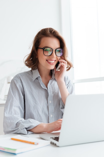 Ritratto del primo piano di giovane donna felice in camicia a strisce che parla sullo smartphone mentre per mezzo del computer portatile a casa