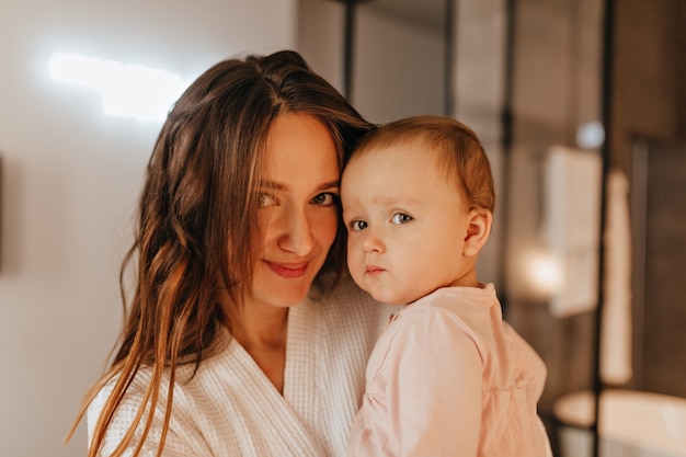 Ritratto del primo piano di giovane donna sorridente che tiene piccola figlia. Il bambino e la madre stanno proponendo in appartamento.