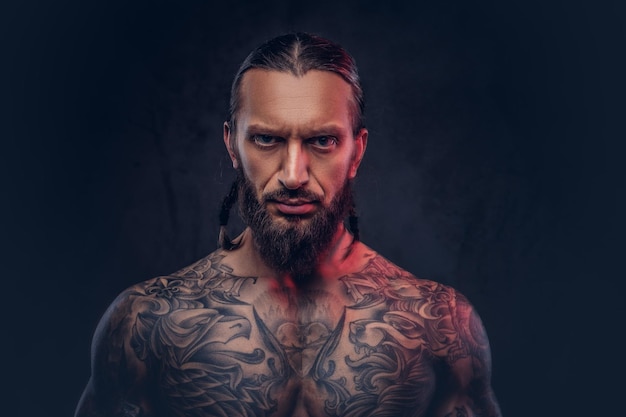 Ritratto del primo piano di un maschio tatuato barbuto muscoloso con un taglio di capelli elegante. Isolato su uno sfondo scuro.