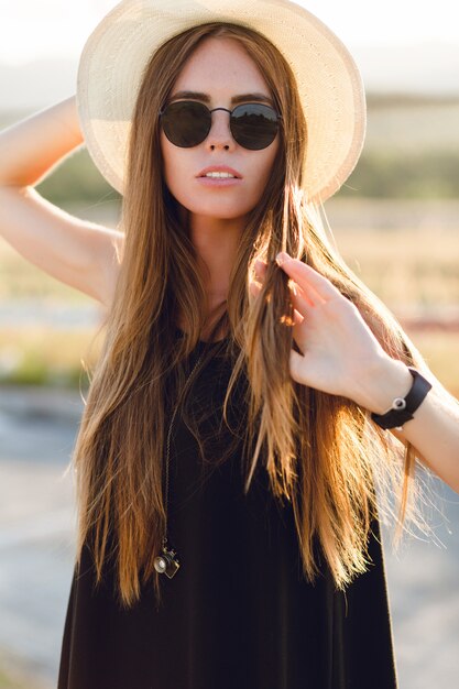 Ritratto del primo piano di una bella ragazza con lunghi capelli scuri che indossa cappello di paglia, occhiali da sole scuri. Gioca con i suoi capelli sotto i caldi raggi del sole al tramonto