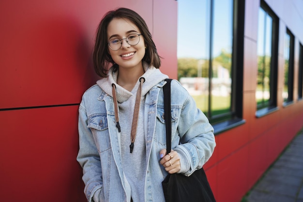 Ritratto del primo piano di una donna 20s allegra e piuttosto giovane con gli occhiali, studente o dipendente, sorridente, fotocamera, felice, borsa, tote bag, magro muro di mattoni rossi del campus, godendo del tempo libero