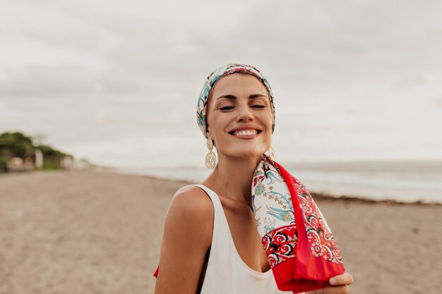 Ritratto del primo piano esterno della donna affascinante sorridente con scialle sulla testa e costume da bagno bianco sulla lunga spiaggia sabbiosa