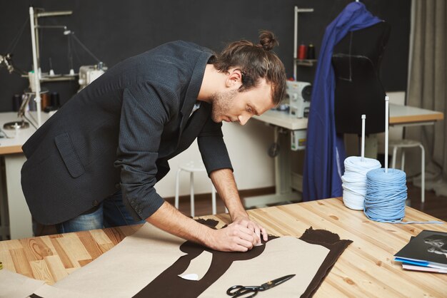 Ritratto del progettista di vestiti maschio adulto bello virile con l'acconciatura alla moda in vestito nero che taglia le parti del vestito futuro da tessuto. L'uomo si è concentrato sul lavoro.