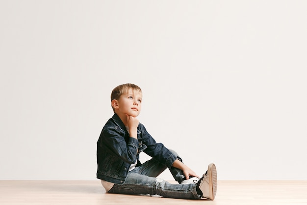 ritratto del ragazzino sveglio in vestiti di jeans alla moda che guarda l'obbiettivo contro il muro bianco dello studio. Concetto di moda per bambini