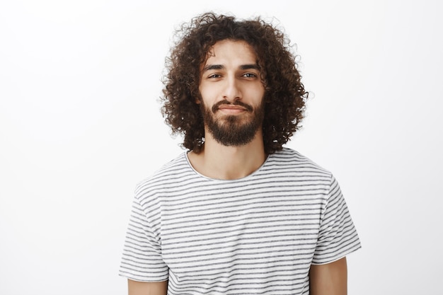 Ritratto del ragazzo creativo fiducioso bello con capelli ricci scuri, in piedi con un leggero sorriso e un'espressione sicura di sé