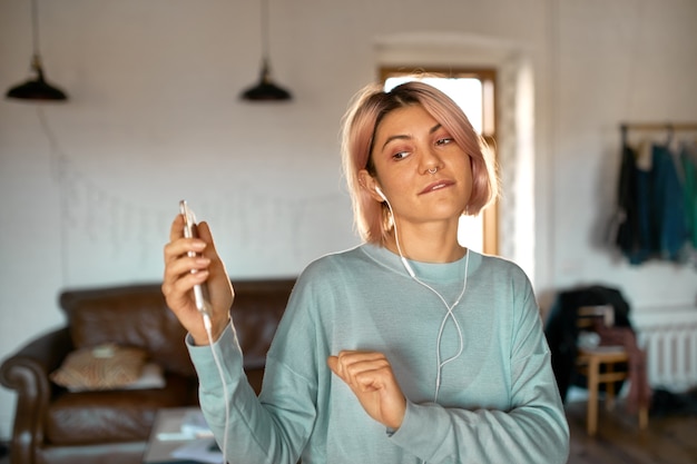 Ritratto dell'interno della ragazza teenager alla moda con capelli rosati che balla alla musica pop utilizzando auricolari e cellulare.