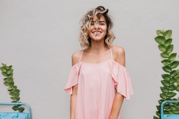 Ritratto dell'interno della signora dai capelli corti spensierata con la pelle abbronzata. sorridente ragazza accattivante in abito rosa in posa con piante verdi.