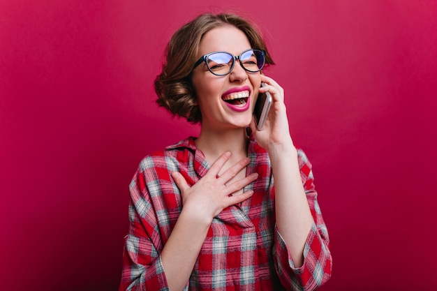 Ritratto dell'interno di ridere splendida ragazza con gli occhiali parlando al telefono sulla parete bordeaux. Foto di entusiasta signora bianca in camicia a scacchi che tiene smartphone e sorridente.