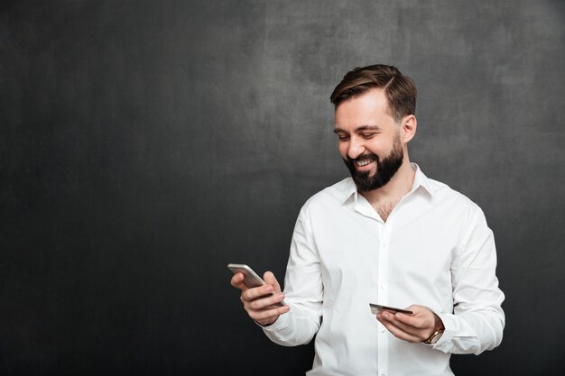 Ritratto dell'uomo allegro che effettua pagamento online in Internet facendo uso del telefono cellulare e della carta di credito, isolato sopra grigio scuro