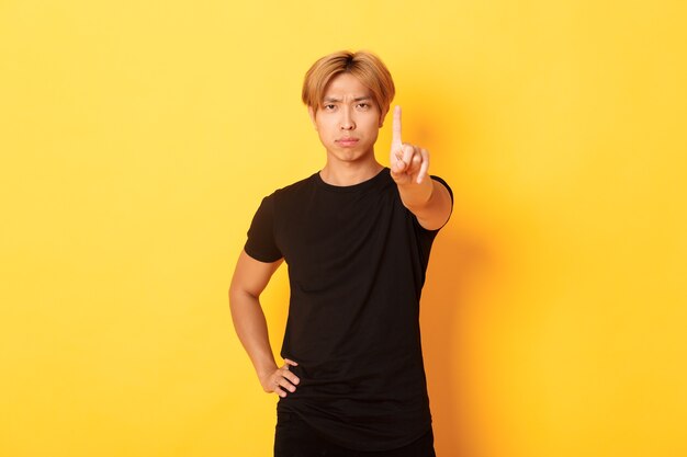 Ritratto dell'uomo asiatico deluso dall'aspetto serio che agita il dito per rimproverare qualcuno, muro giallo in piedi
