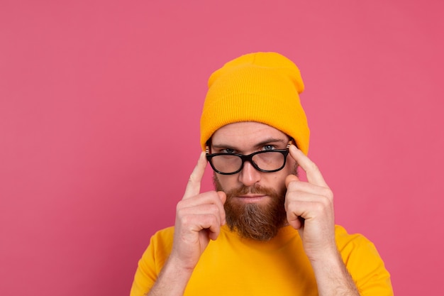 Ritratto dell'uomo barbuto europeo bello alla moda in cappello e occhiali gialli casuali della camicia sul colore rosa