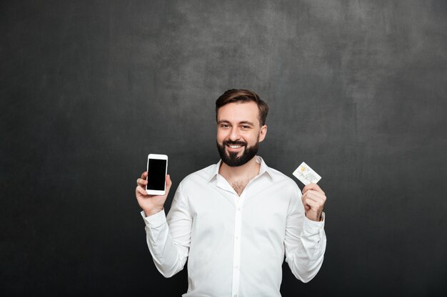 Ritratto dell'uomo castana che posa sulla macchina fotografica facendo uso dello smartphone e della carta di credito per acquisto online, isolato sopra grigio scuro