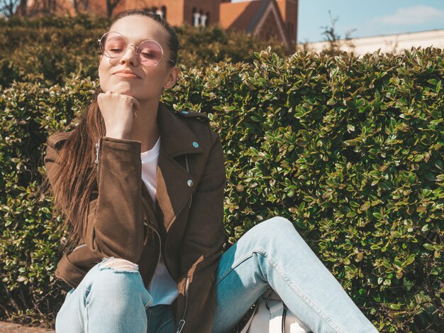 Ritratto della bellissima modella bruna sorridente vestita in abiti hipster estate giacca e jeans Ragazza alla moda, seduto sulla panchina in strada