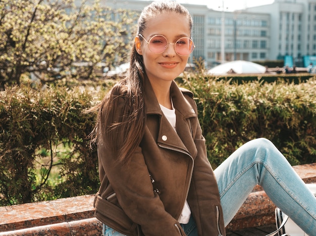 Ritratto della bellissima modella bruna sorridente vestita in abiti hipster giacca e jeans alla moda Ragazza alla moda, seduto sulla panchina in strada Donna divertente e positiva in occhiali da sole