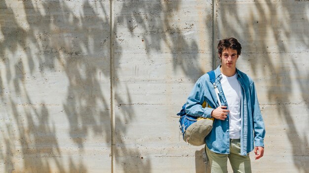 Ritratto della borsa di trasporto dell'uomo bello che esamina macchina fotografica che sta contro il muro di cemento