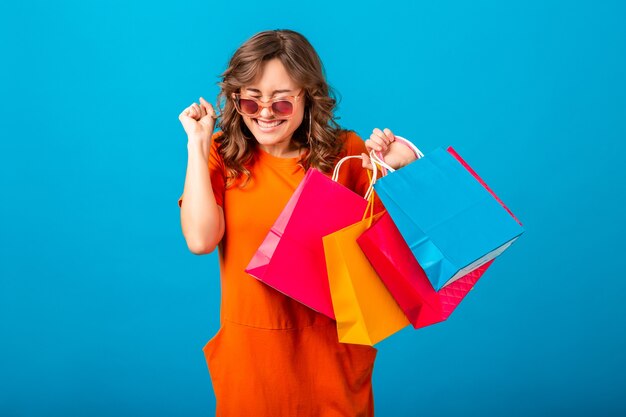 Ritratto della donna alla moda sorridente attraente eccitata dello shopping in vestito alla moda arancione che tiene i sacchetti della spesa sul fondo blu dello studio isolato