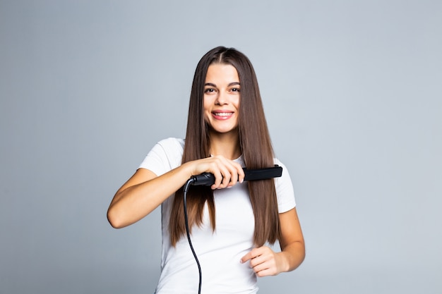 Ritratto della donna allegra che usando raddrizzatore per i suoi capelli ricci che preparano per la pettinatura facile comoda di festa della data di evento isolata su gray