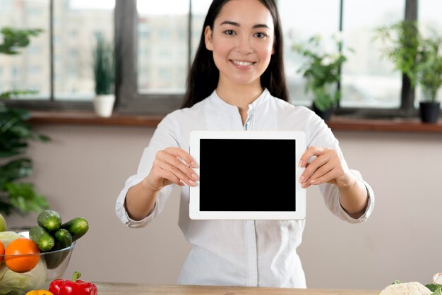 Ritratto della donna asiatica che mostra compressa digitale dello schermo in bianco in cucina