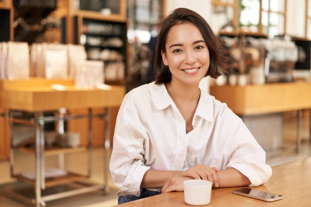 Ritratto della donna asiatica sicura che si siede nello smartphone del caffè e nel caffè sul tavolo smili della donna di affari