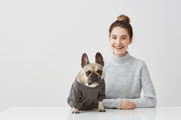 Ritratto della donna castana sorridente con capelli legati nell'ufficio di lavoro di topknot a casa. Free lance femminili che si siedono al tavolo in officina in compagnia del cane. Concetto di amicizia