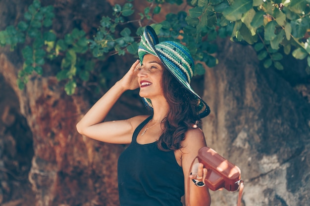 Ritratto della donna che sta e che tiene il suoi cappello e camerand in camicia nera alla spiaggia durante il giorno.