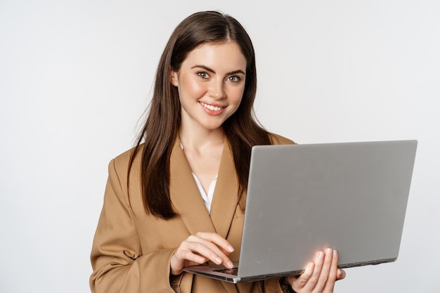 Ritratto della donna corporativa che lavora con il computer portatile che sorride e che osserva il fondo bianco assertivo
