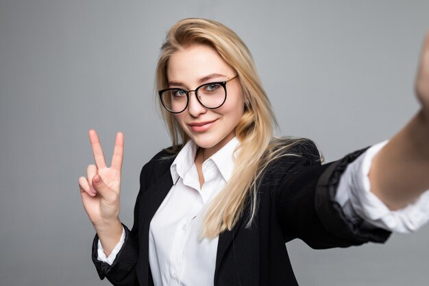 Ritratto della donna di affari che fa selfie facendo uso dello Smart Phone e di Internet, gesturing simbolo di pace, avendo video chiamata online sopra la parete grigia