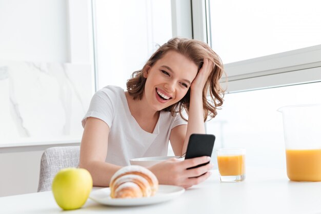 Ritratto della donna di risata in maglietta bianca che controlla le notizie sul telefono cellulare mentre sedendosi al tavolo da cucina