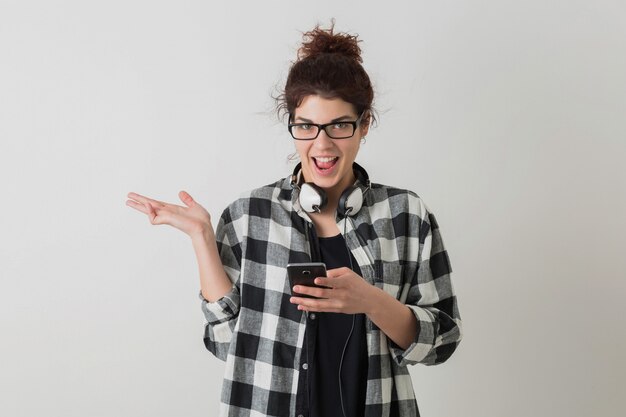 Ritratto della donna graziosa sorridente dei giovani pantaloni a vita bassa nella posa d'uso di vetro della camicia a quadretti isolata, tenendo Smart Phone, fronte divertente pazzo