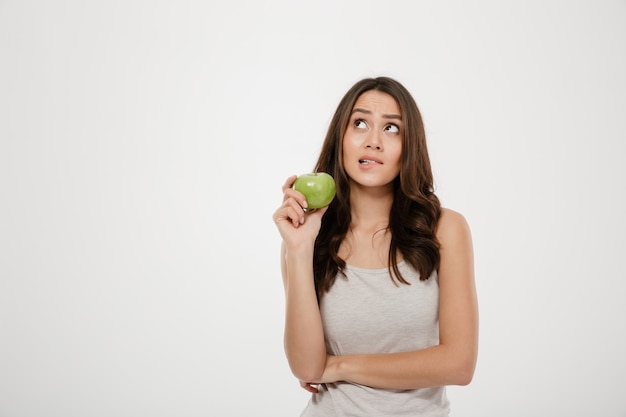 Ritratto della donna imbarazzata che guarda verso l'alto tenendo mela fresca verde, pensando all'alimento sano isolato sopra bianco