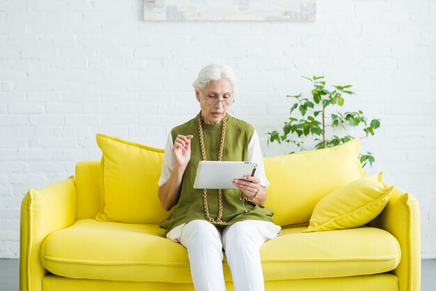 Ritratto della donna senior che si siede sul sofà giallo che esamina compressa digitale