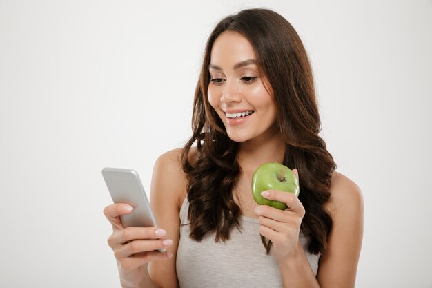 Ritratto della donna soddisfatta che per mezzo dello smartphone d'argento mentre mangiando mela verde fresca, isolato sopra la parete bianca