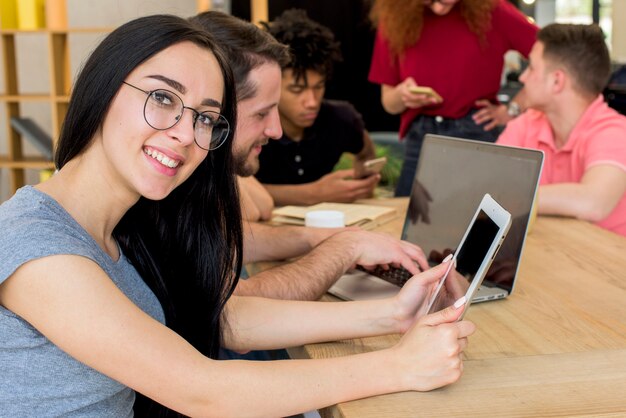 Ritratto della donna sorridente che tiene compressa digitale che esamina macchina fotografica mentre sedendosi accanto ai suoi amici facendo uso degli aggeggi elettronici e del libro sullo scrittorio di legno