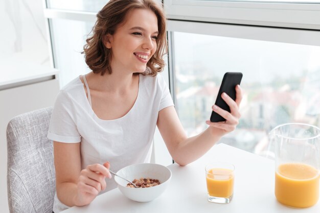 Ritratto della donna sorridente sveglia in maglietta bianca che chiacchiera sullo smartphone mentre sedendosi e mangiando i fiocchi di granturco al tavolo da cucina