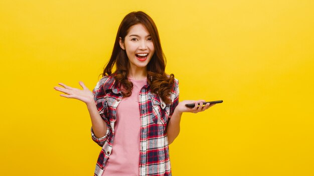 Ritratto della femmina asiatica sorpresa che per mezzo del telefono cellulare con l'espressione positiva, vestita in abbigliamento casual ed esaminando macchina fotografica sopra la parete gialla. La donna felice adorabile felice si rallegra del successo.