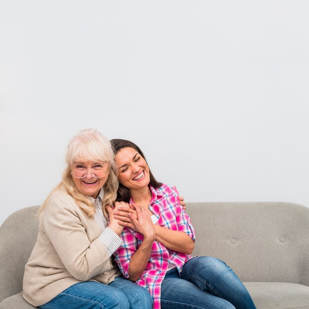 Ritratto della madre e sua figlia che si siedono insieme sul sofà contro la parete bianca