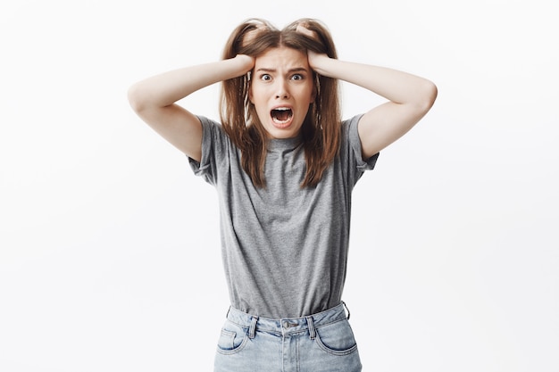 Ritratto della ragazza caucasica divertente del brunette in vestiti grigi che tengono capelli con le mani, con la bocca aperta e l'espressione spaventata. Donna che grida dopo aver perso il suo telefono cellulare.